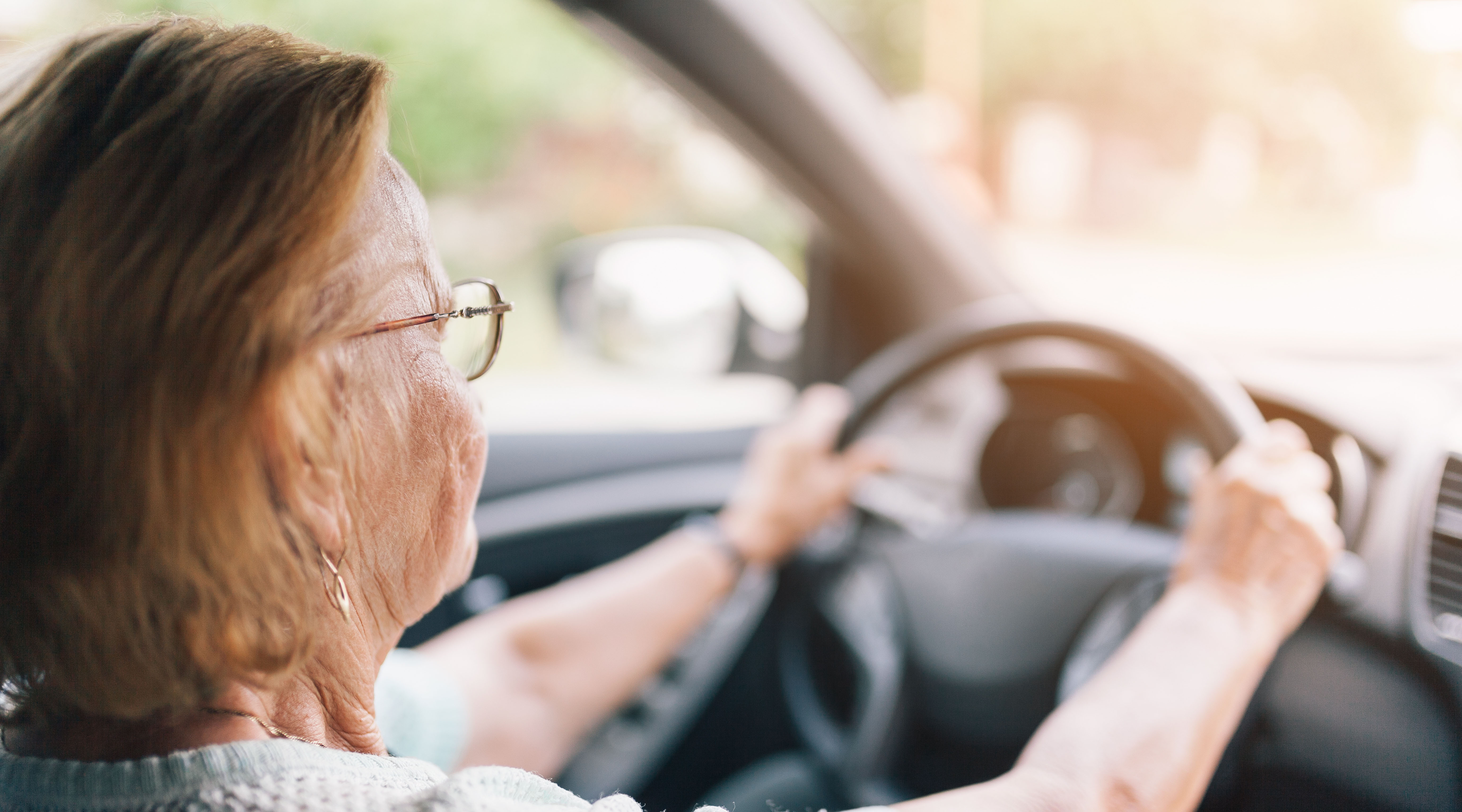 Perspective over the shoulder shot of an older lady with glasses holding the steering wheel.